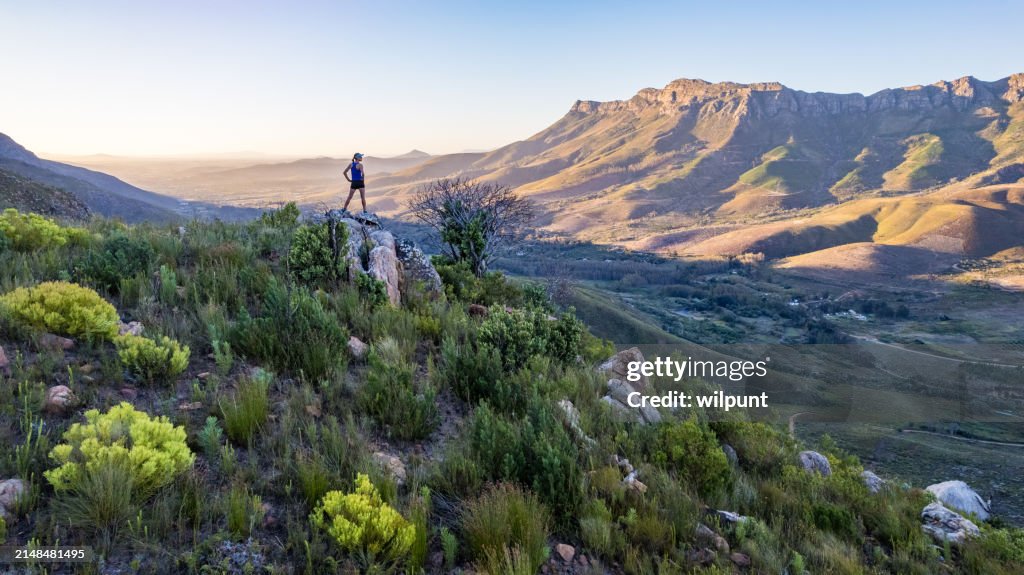 Silueta femenina del corredor del sendero de pie en el pico de la montaña mirando hacia fuera de la cordillera al atardecer