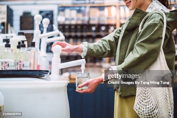 sustainable choices: a young asian woman refilling a glass jar with biodegradable cleaning product in zero-waste store - cleaning equipment stock pictures, royalty-free photos & images