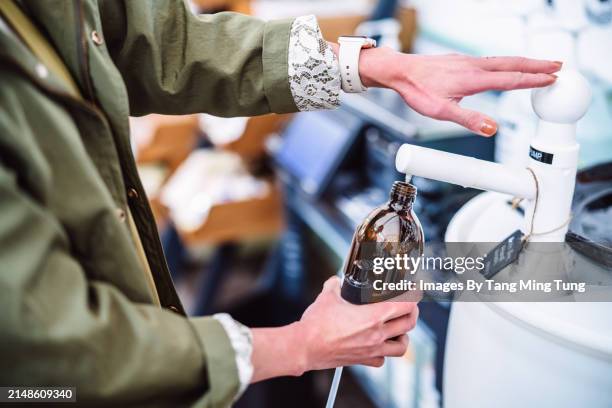 sustainable choices: a young female shopper refilling a reusable glass bottle with body care product in zero-waste store - grünes wirtschaften stock-fotos und bilder
