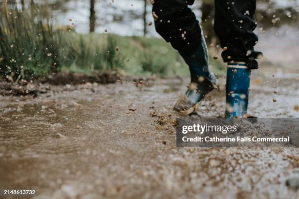 a child creates splashes and ripples as he wades through a muddle puddle wearing blue wellington boots - pluie diluvienne photos et images de collection