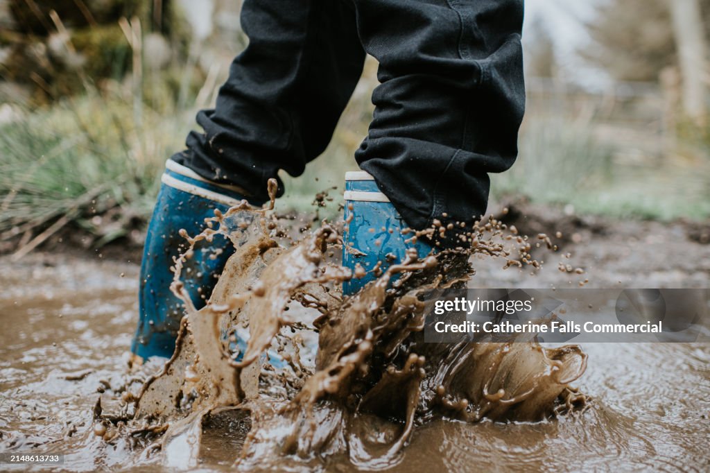 A child creates splashes and ripples as he wades through a muddle puddle wearing blue wellington boots