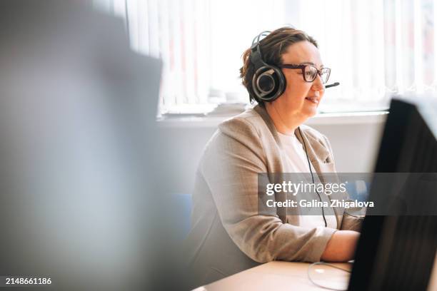 smiling middle aged brunette woman in headset with headphones and microphone working at computer in the office - telemarketing stock pictures, royalty-free photos & images