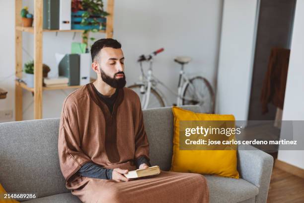 a serene young adult with a beard meditates while holding a book, seated on a modern couch with vibrant cushions, exuding calm in a well-lit living space. - exuding stock pictures, royalty-free photos & images