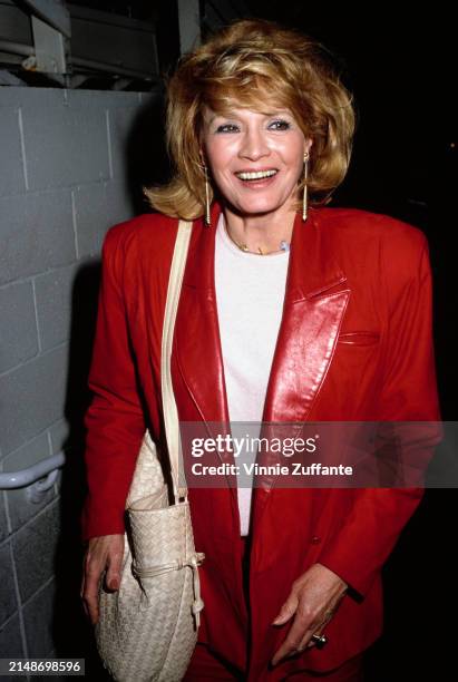 American actress Angie Dickinson, wearing a red jacket over a white crew neck top, with a white shoulder bag over her right shoulder, United States,...