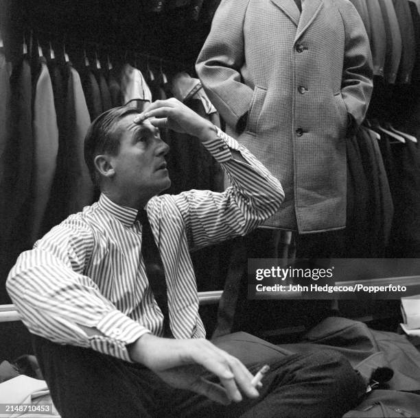 English fashion designer Hardy Amies seated on the floor of a men's outfitters shop as he dresses a male model with a selection of coats of varying...