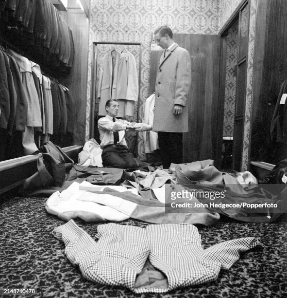 English fashion designer Hardy Amies kneels on the floor of a men's outfitters shop as he dresses a male model with a selection of coats of varying...