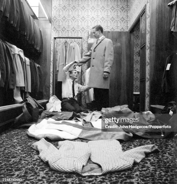 English fashion designer Hardy Amies kneels on the floor of a men's outfitters shop as he dresses a male model with a selection of coats of varying...