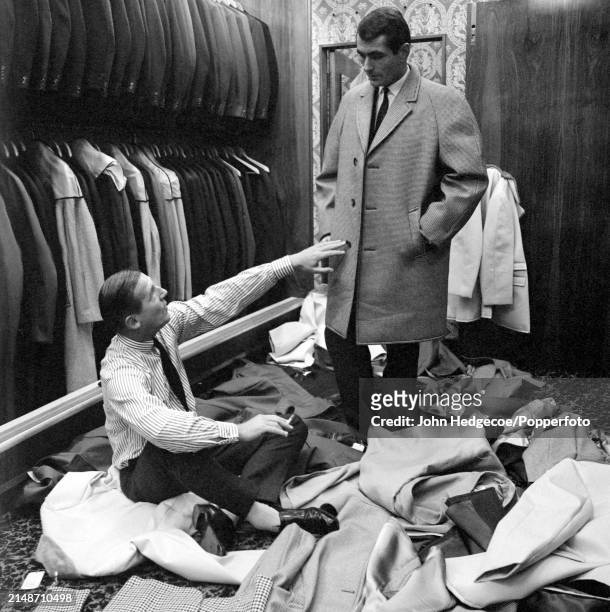 English fashion designer Hardy Amies seated on the floor of a men's outfitters shop as he dresses a male model with a selection of coats of varying...