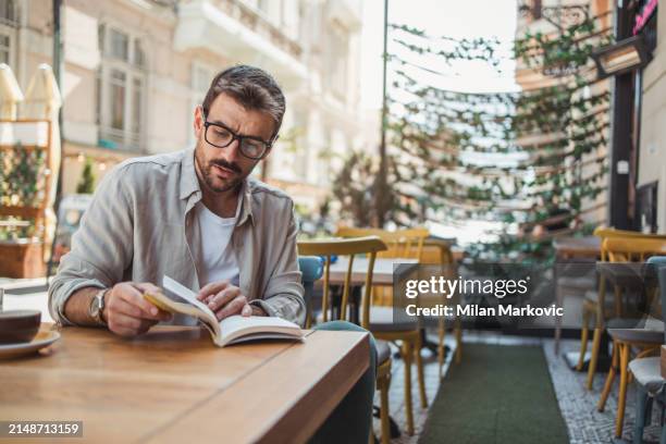 un joven está leyendo un libro mientras está sentado en el jardín de un café - un solo hombre fotografías e imágenes de stock