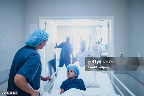 young girl pushed into bright surgery room with medical staff in a hospital mother assisting - black child hospital bed stock pictures, royalty-free photos & images