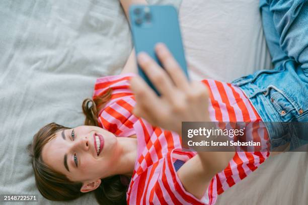 happy young woman using smartphone lying on the bed at home - mit dem gesicht nach unten stock-fotos und bilder