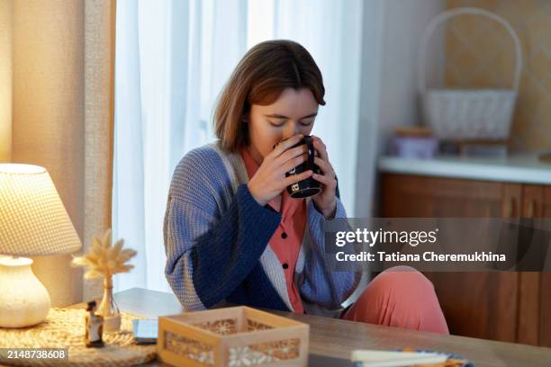 young woman with a cold drinking hot tea from a mug while recovering in kitchen room at home - cold virus stock pictures, royalty-free photos & images