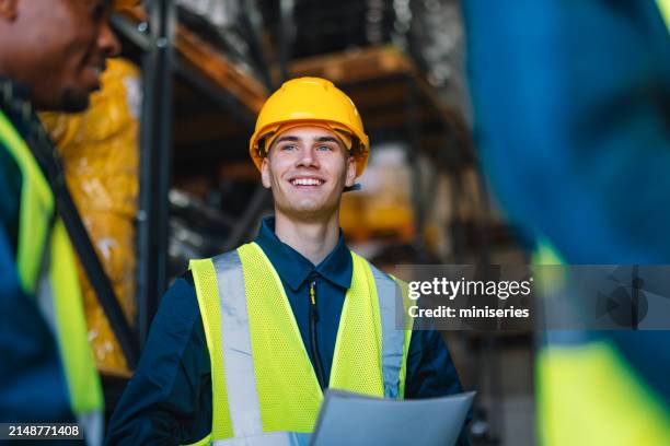 un joven trabajador de almacén feliz con un casco amarillo y un chaleco de seguridad se encuentra interactuando con sus colegas en un bullicioso entorno industrial. - material reflector de seguridad fotografías e imágenes de stock