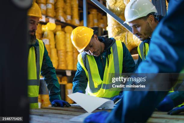 un groupe d’employés d’entrepôt en tenue de sécurité se rassemble autour d’un document. ils discutent des protocoles de sécurité dans un environnement industriel entouré de matériaux. - obéissance photos et images de collection