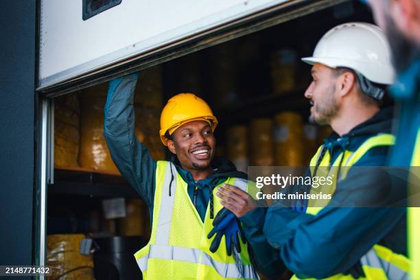 two industrial warehouse workers in safety helmets and reflective vests engage joyfully near the storage area, exemplifying teamwork and workplace safety. - loading dock stock pictures, royalty-free photos & images