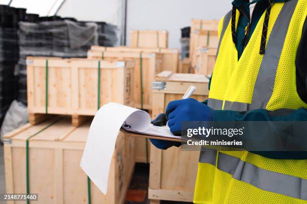 a focused warehouse worker wearing high-visibility safety gear audits boxes in a logistics warehouse. he notes details on a clipboard, ensuring efficiency and accuracy in inventory management. - regels stockfoto's en -beelden