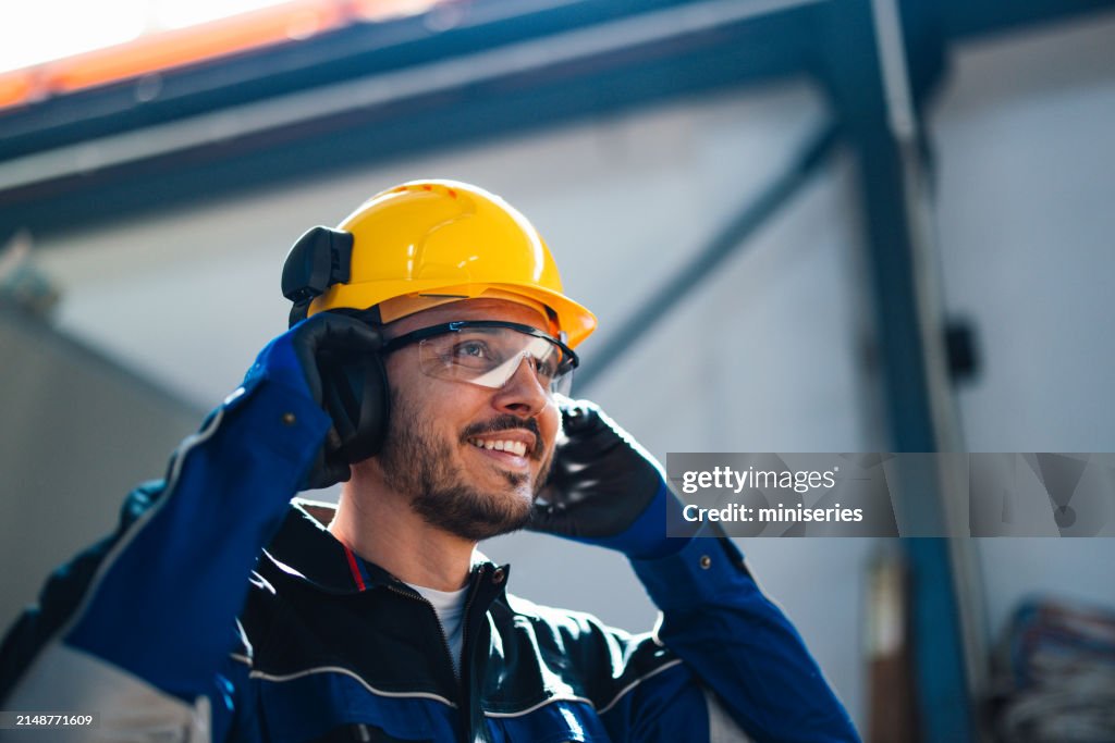 Ein fröhlicher männlicher Arbeiter mit Schutzhelm und Schutzausrüstung spricht in einer geschäftigen Fabrikumgebung über ein Headset und betont die Sicherheit und Produktivität der Branche.