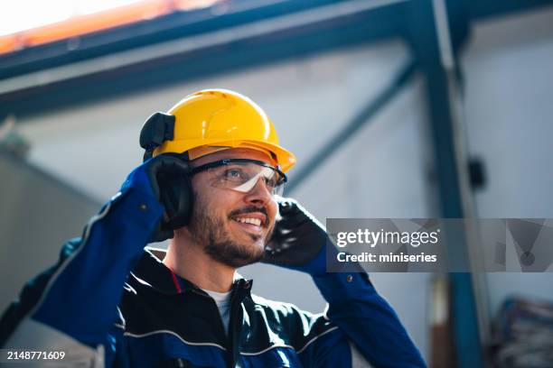 un alegre trabajador con casco de seguridad y equipo de protección habla por auriculares en un bullicioso entorno de fábrica, haciendo hincapié en la seguridad y la productividad de la industria. - casco de trabajo fotografías e imágenes de stock