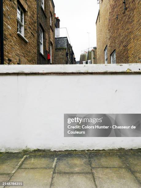 concrete wall painted white behind which we can see the rear facades in weathered bricks of houses and residential buildings. dirty cement sidewalk.
london, england, united kingdom.
sunlight. natural colors.
no people. - pavement stock pictures, royalty-free photos & images