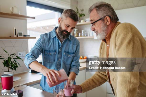 senior father and mature son making fruit smoothie as snack, having fun in kitchen. handsome man pouring, serving finished smoothie into glasses. - gelenkrheumatismus stock-fotos und bilder