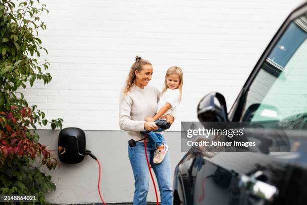 smiling woman carrying daughter while walking towards electric car with charger - elektrische auto stockfoto's en -beelden