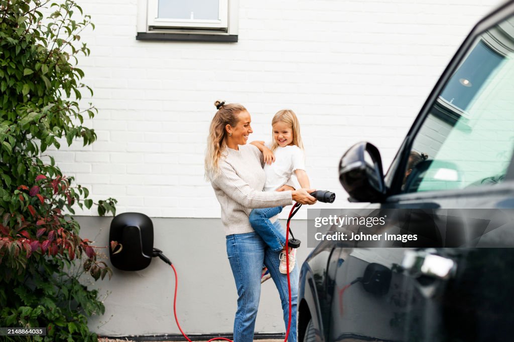 Happy woman carrying girl while walking towards electric car with charger