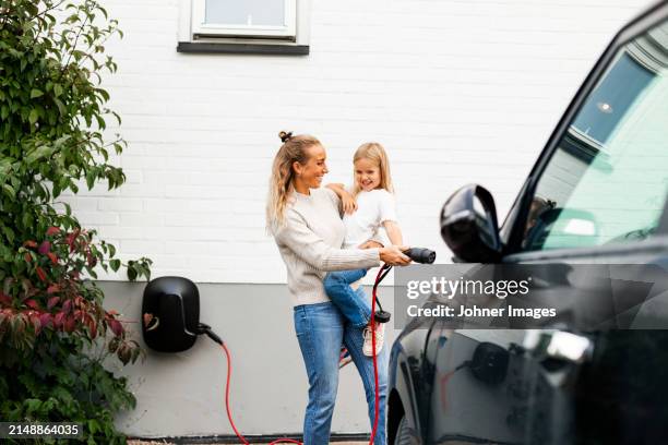 Happy Woman Carrying Girl While Walking Towards Electric Car With Charger, Foto de stock