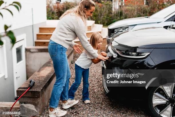 full length of mother assisting daughter with charging electric car at front yard - elektrische auto stockfoto's en -beelden