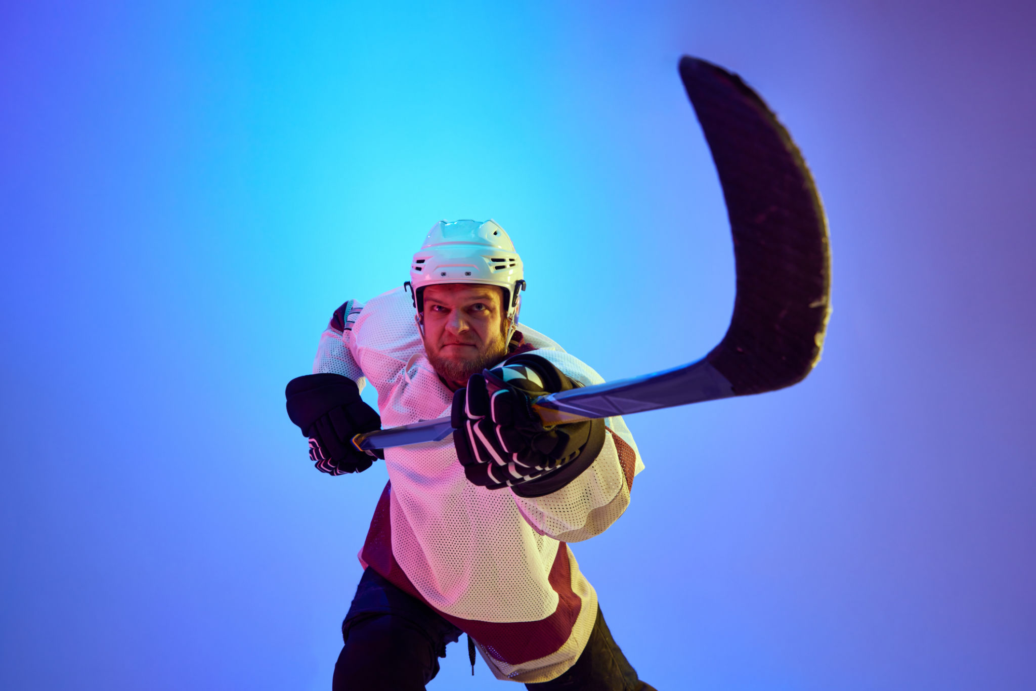 Competitive, motivated man, hockey player posing with stick, in uniform and helmet against gradient blue background in neon light Competitive, motivated man, hockey player posing with stick, in uniform and helmet against gradient blue background in neon light