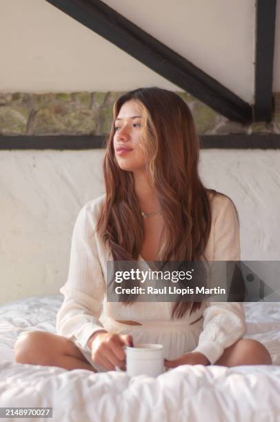 a woman sits on a bed, basking in sunlight, holding a coffee cup, exuding a sense of calm. - exuding stock pictures, royalty-free photos & images