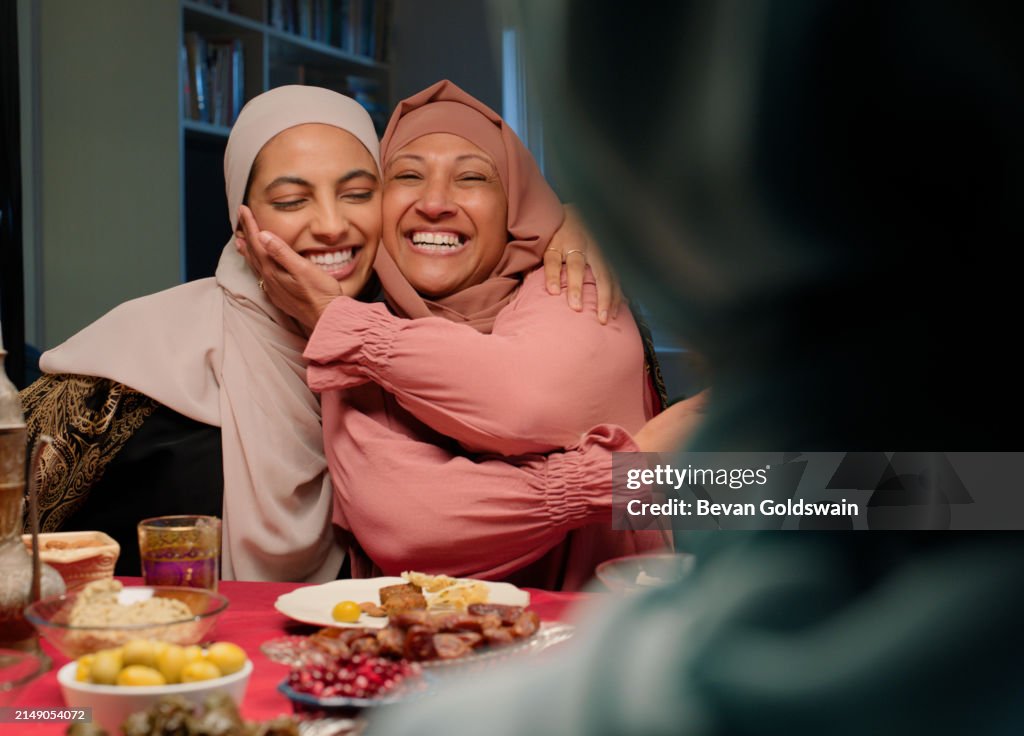 Islam, Umarmung und Essen für die Ramadan-Feier, muslimische Menschen am Esstisch, die gemeinsam essen, lachen und lächeln. Frauen, Umarmung und Familie mit Kultur, Fest oder fröhlichem Fest von Eid al Fitr zu Hause