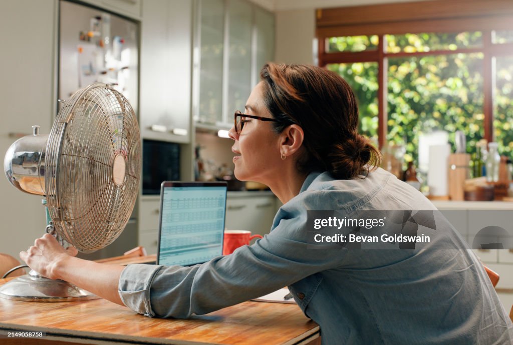 Frau, Ventilator und Laptop zu Hause mit Hitzewelle als freiberufliches Texten für Remote-Arbeit, E-Mail oder Küche. Weibliche Person, reif und Menopause bei heißem Wetter oder Wohnungsgerät, kühler Luft oder Sommer