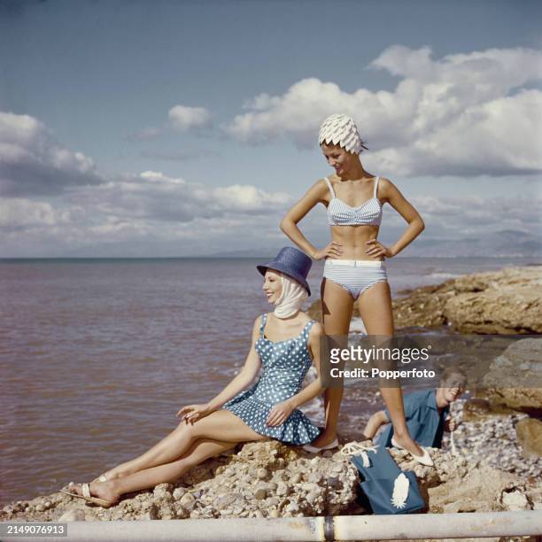 Vacation scene of two female fashion models wearing swimsuits, from left, a pale blue and white polka dot skirted swimming costume with head scarf...