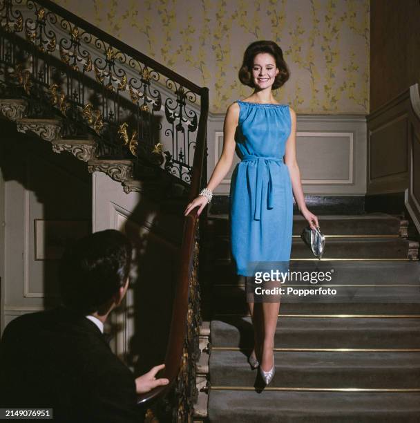 Female fashion model, wearing a blue sleeveless dress with gathered neckline and tie waist, walks down a grand staircase to meet a man standing at...