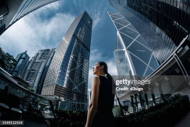 low angle shot of successful young asian businesswoman standing against contemporary corporate skyscrapers. looking up to sky with confidence. female leadership and empowerment. woman in business. determined to success - grand angle photos et images de collection