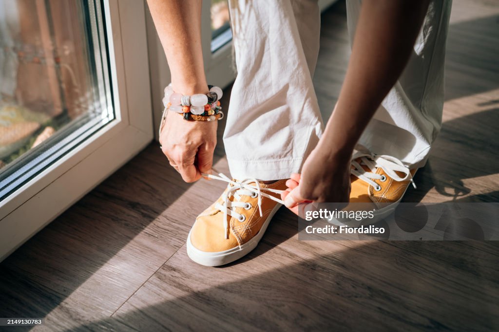 A woman in white trousers tying her shoelaces on a yellow sneaker. The process of dressing and putting on shoes