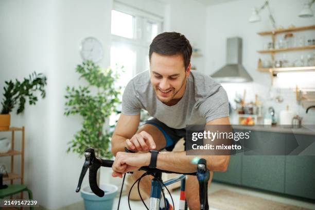 young athletic man checking pulse on smart watch while riding bicycle in a domestic room at home - home workout stock pictures, royalty-free photos & images