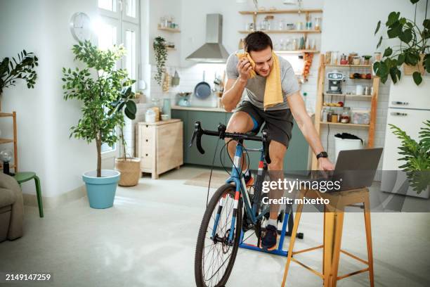 handsome athletic man riding exercise bicycle at home while using laptop - cyclist stock pictures, royalty-free photos & images