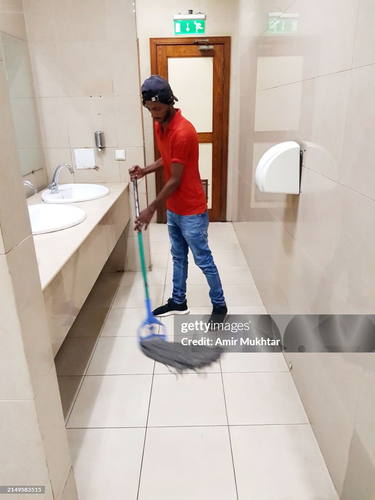 One young restroom attendant cleaning and sweeping the tile floor with broom or mop of office washroom.