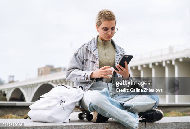 teenage girl with backpack with paper cup of coffee in her hands using mobile phone sitting on skateboard at the city - kort haar stockfoto's en -beelden