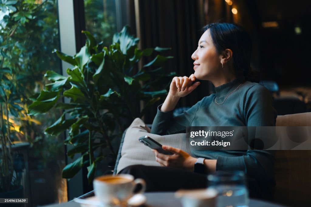 Confident young Asian woman with smartphone looking out through window while sitting in a cafe having coffee. Making a personal financial plans and investment decisions. Wealth management. Business, banking, finance and investment concept