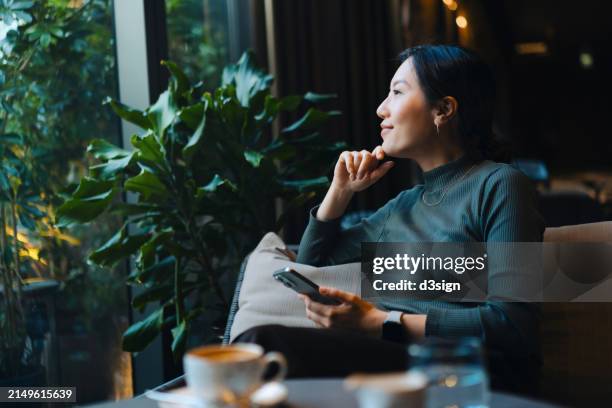 confident young asian woman with smartphone looking out through window while sitting in a cafe having coffee. making a personal financial plans and investment decisions. wealth management. business, banking, finance and investment concept - china oost azië stockfoto's en -beelden