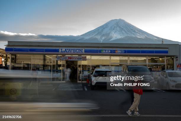 This photo taken on January 1, 2024 shows a tourist posing in front of a convenience store with Mount Fuji in the background, in the town of...