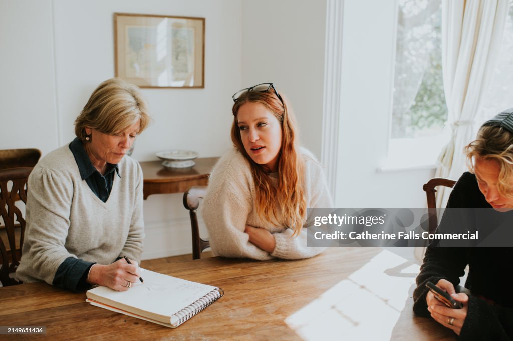 Three woman sit around a dining table in a sunny house, making notes