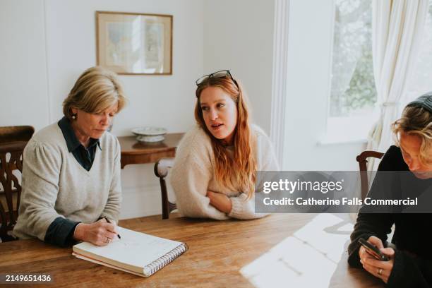 three woman sit around a dining table in a sunny house, making notes - erfenis stockfoto's en -beelden