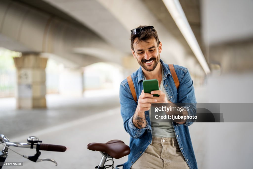 Joven sonriente de pie con su bicicleta y escribiendo en el teléfono