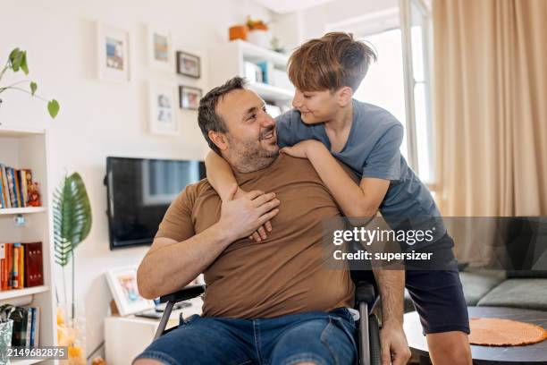 wheelchair-bound dad hugged by his son at home - human relationship stock pictures, royalty-free photos & images