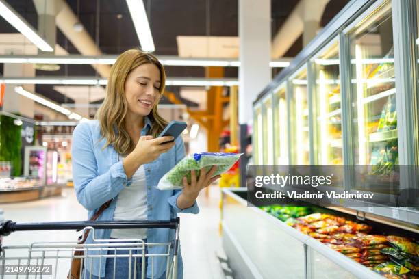 woman looking at smart phone in supermarket - estantería de productos refrigerados fotografías e imágenes de stock