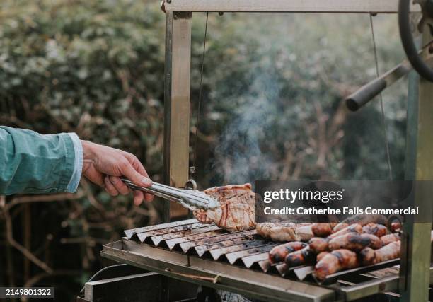 a man uses tongs to turn meat chops on an argentinian ‘asado’ barbecue - costeleta comida imagens e fotografias de stock