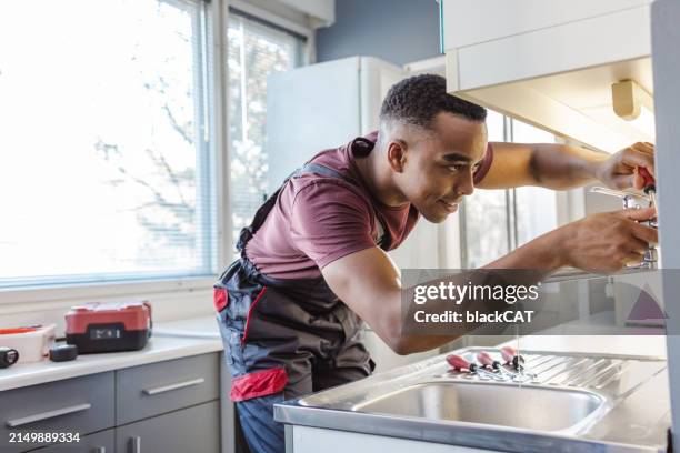 fontanero afroamericano arreglando una fuga en el fregadero de la cocina en la casa del cliente - fontanero fotografías e imágenes de stock